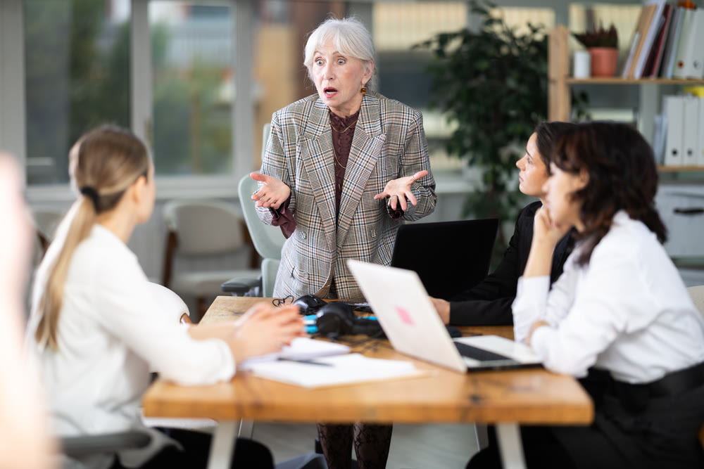 Standing senior woman scold to round table gathering female staff at office