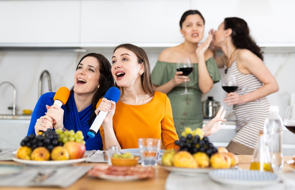 Group of female friends having fun at alcoholic bachelorette party in cozy living room, drinking red wine and singing together at home karaoke