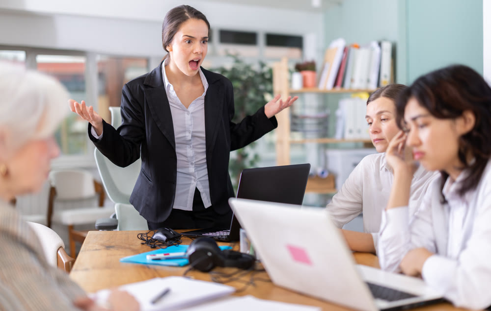 Young female manager standing and scolding colleagues during stressful corporate meeting
