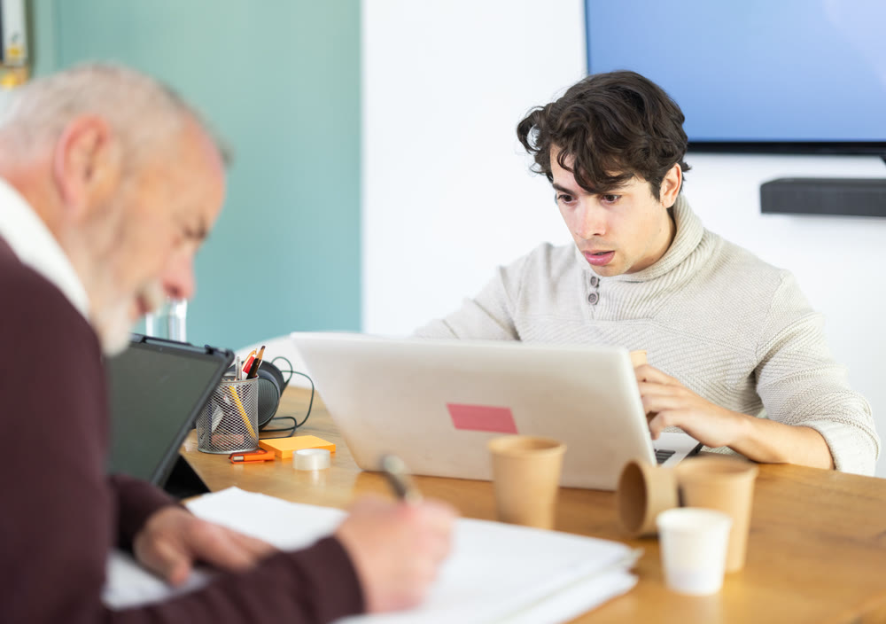 Young man working on laptop, elderly man takes notes on documents