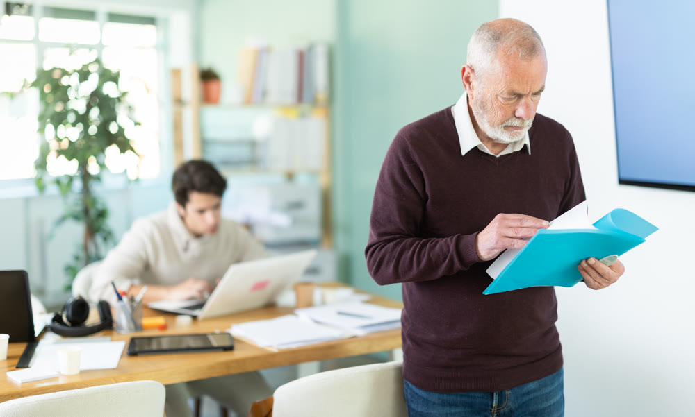 Elderly man reviews documents young man works on laptop in office