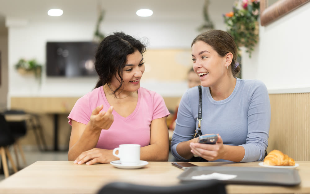 Latin girl is chatting with female friend at table in cafe, showing photos on phone screen