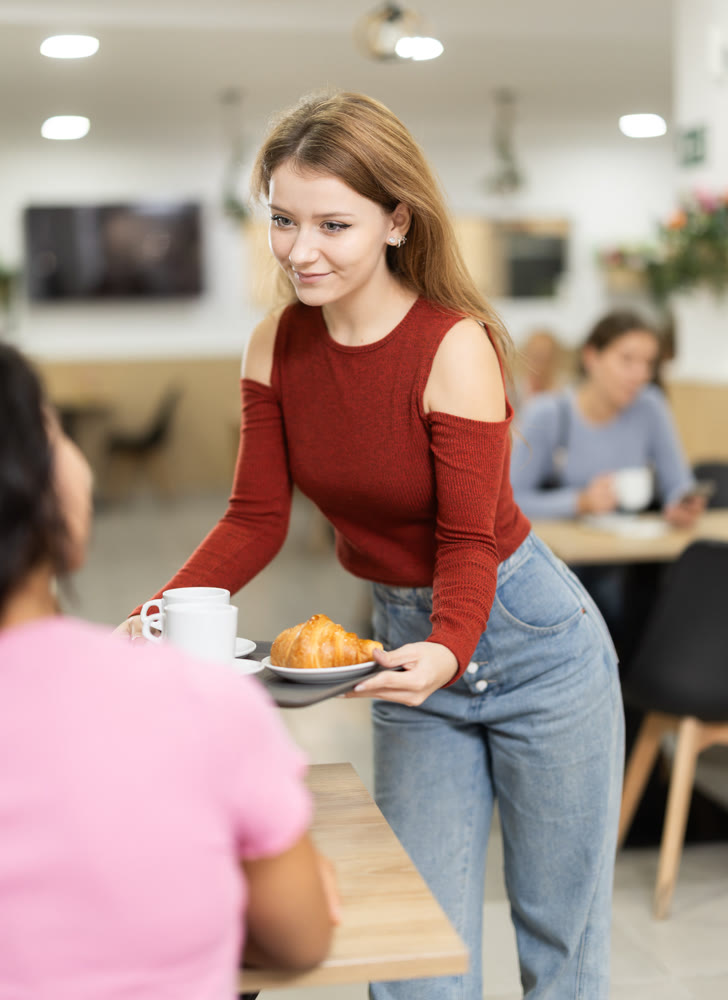 Young woman carries coffee and croissant to table