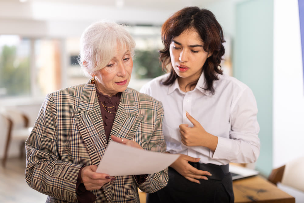Mature woman and young colleague discussing documents while standing in office