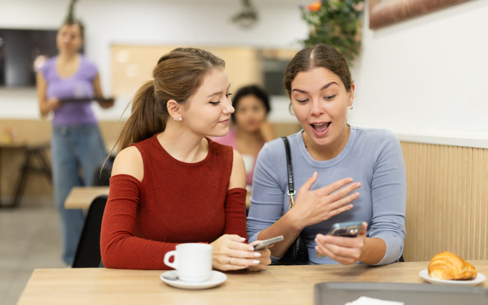 Girl is chatting with female friend at table in cafe, showing photos on phone screen