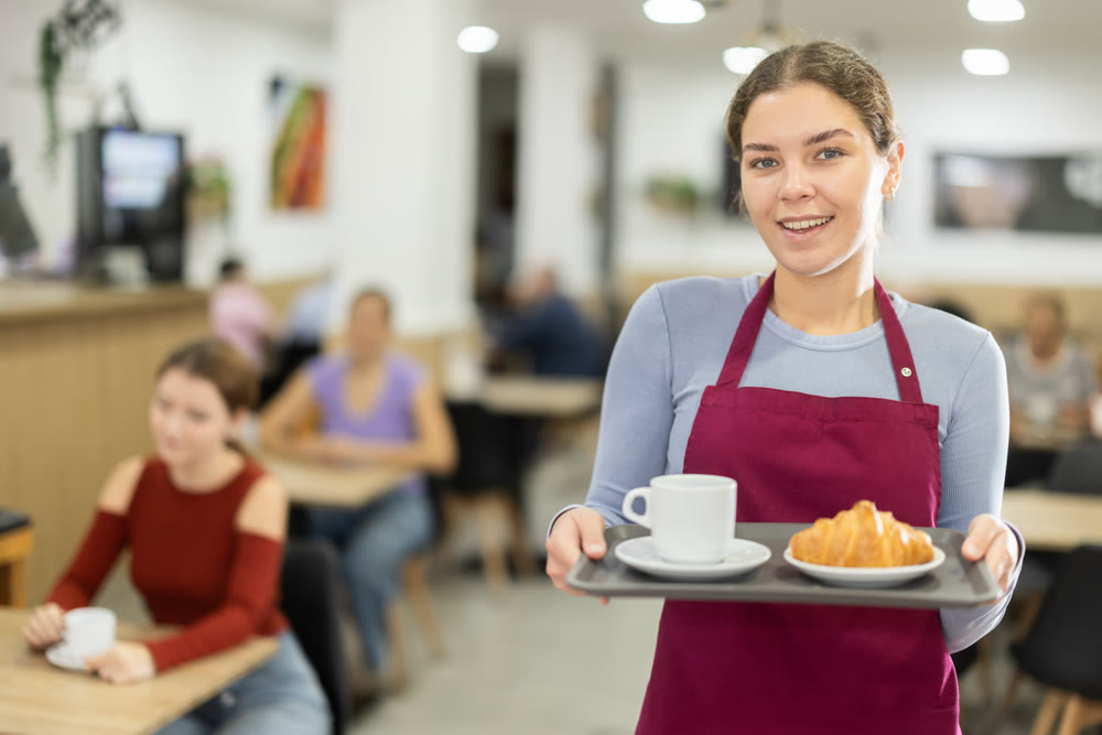 Polite waitress holding tray with coffee and croissant in cafeteria