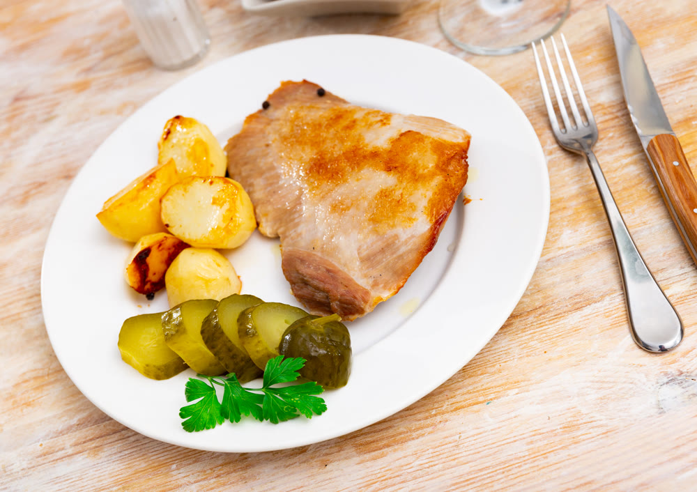 Lunch served on plate - fried pork chop with side dish baked roasted unpeeled potato, and slices of cucumber pickles. Meat is cooked with spice corn black pepper