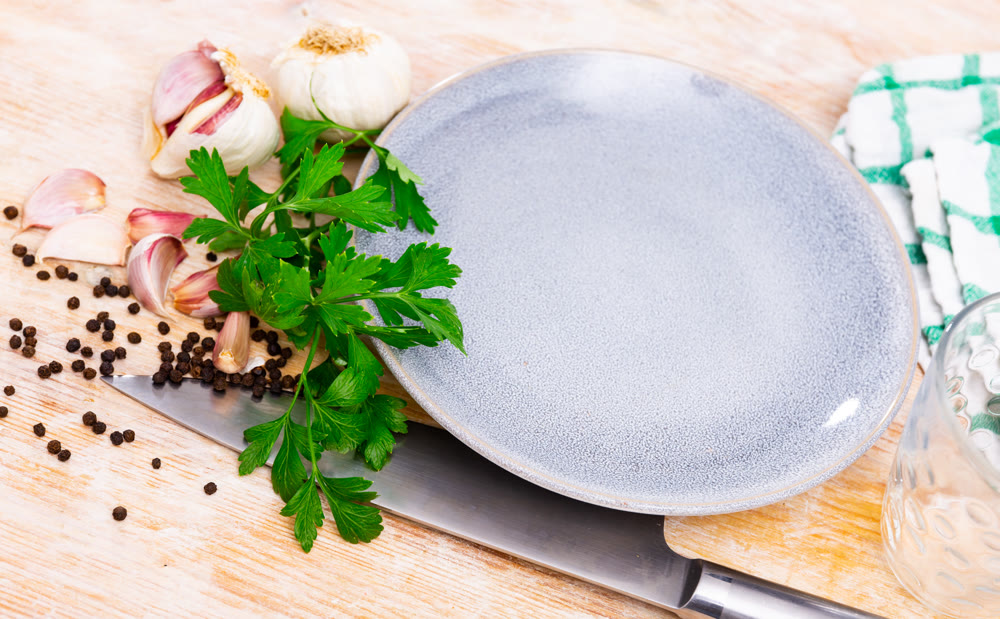 On table there are several cloves of garlic, parsley next to white plate