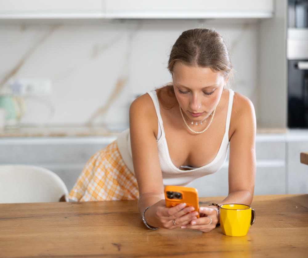 Girl in shorts enjoys drinking coffee or tea and look smartphone in kitchen