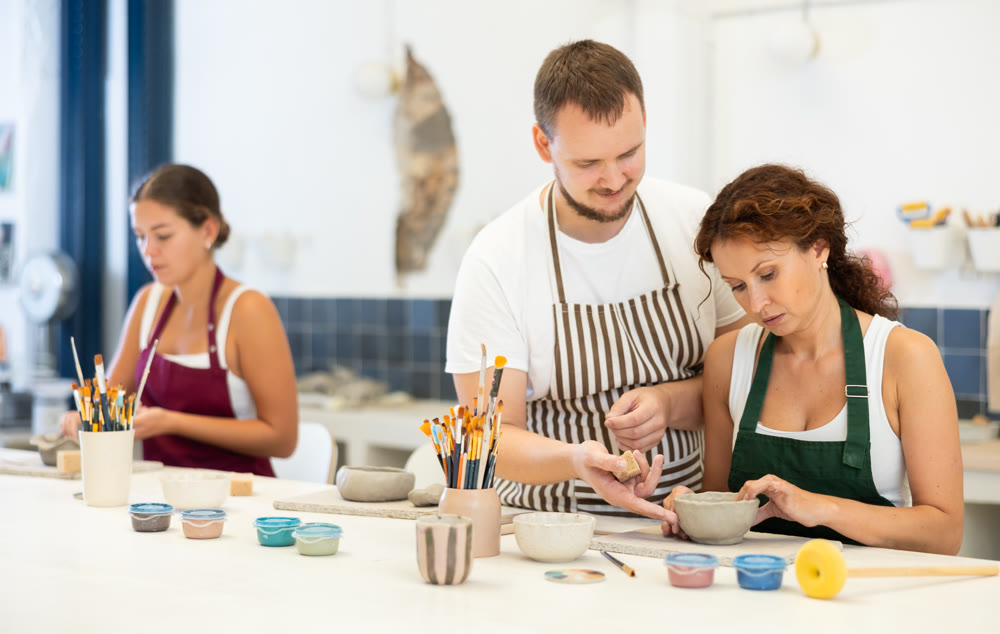 Male pottery teacher helps female students shape clay into plates and other dishes