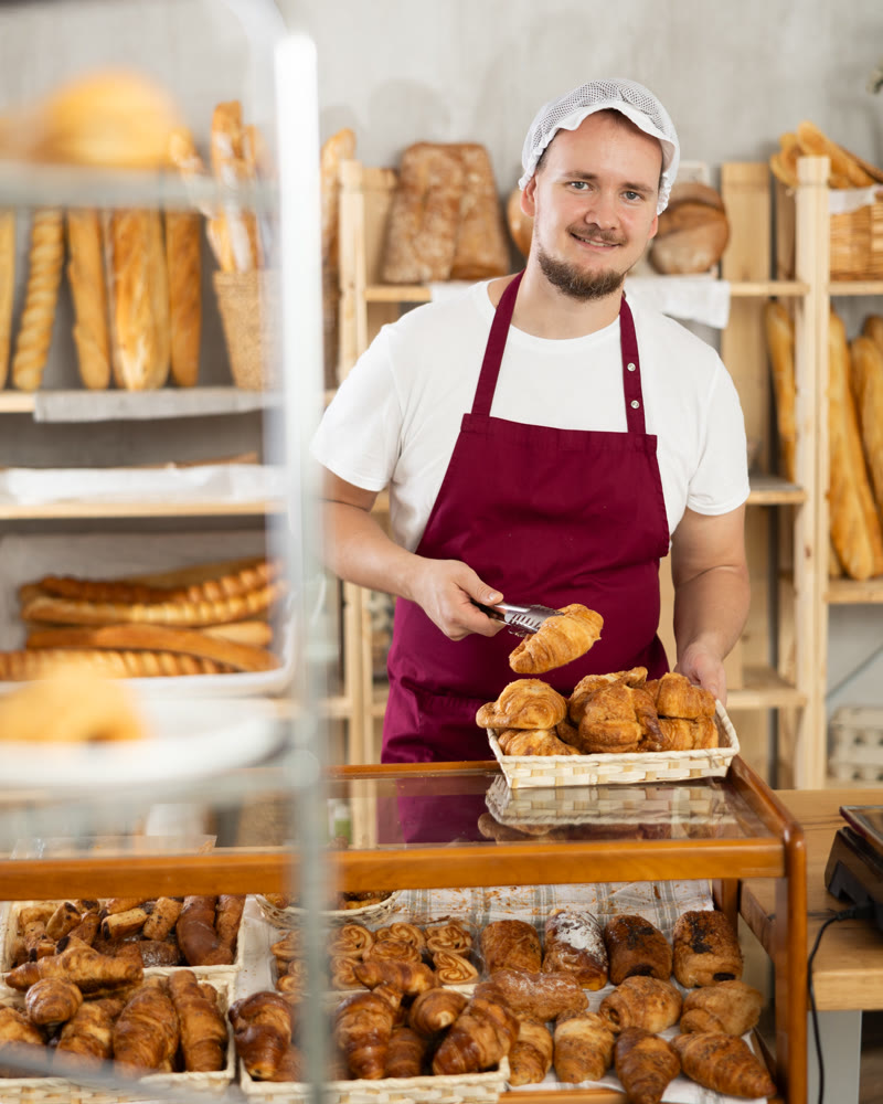 Young man employee puts croissants in window, arranges display of goods at bakery.