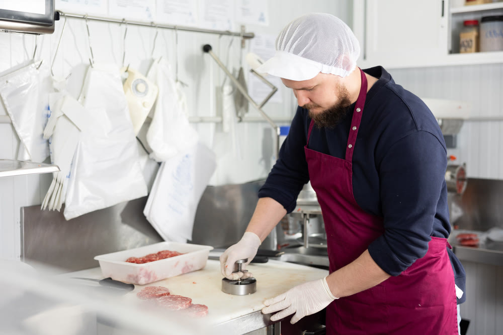 Male food company employee prepares raw meat cutlets using special equipment
