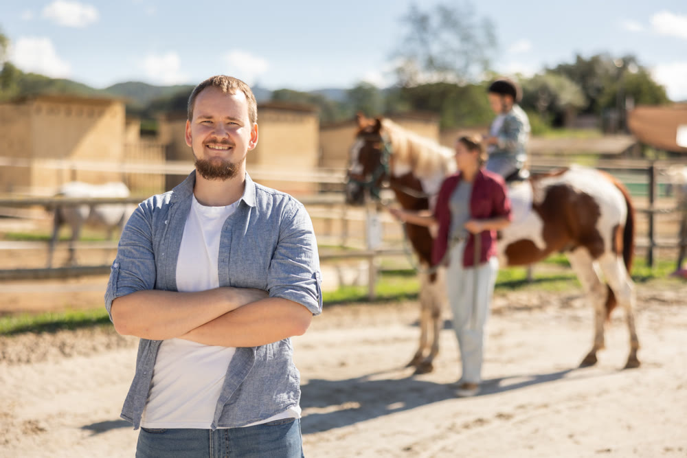 Portrait of happy father in front of horse farm. In background, her wife and son are riding horse
