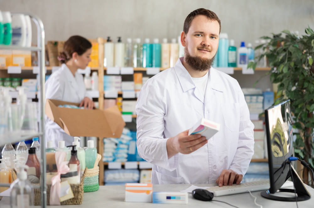 Man pharmacist uses computer at workplace. Girl assistant in background
