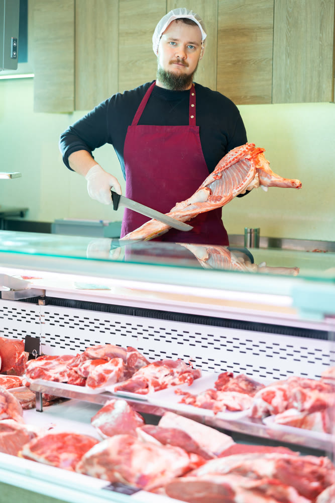 Skilled bearded butcher cutting fresh lamb carcass at counter