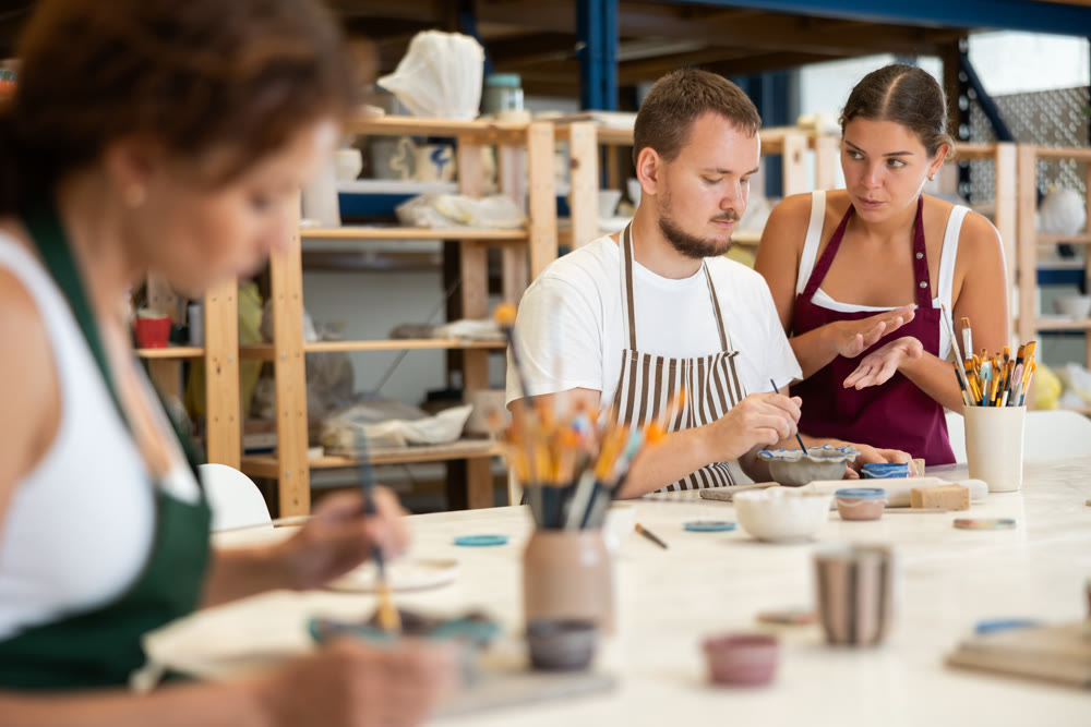 During master class, student paints clay vessel under guidance of female teacher.