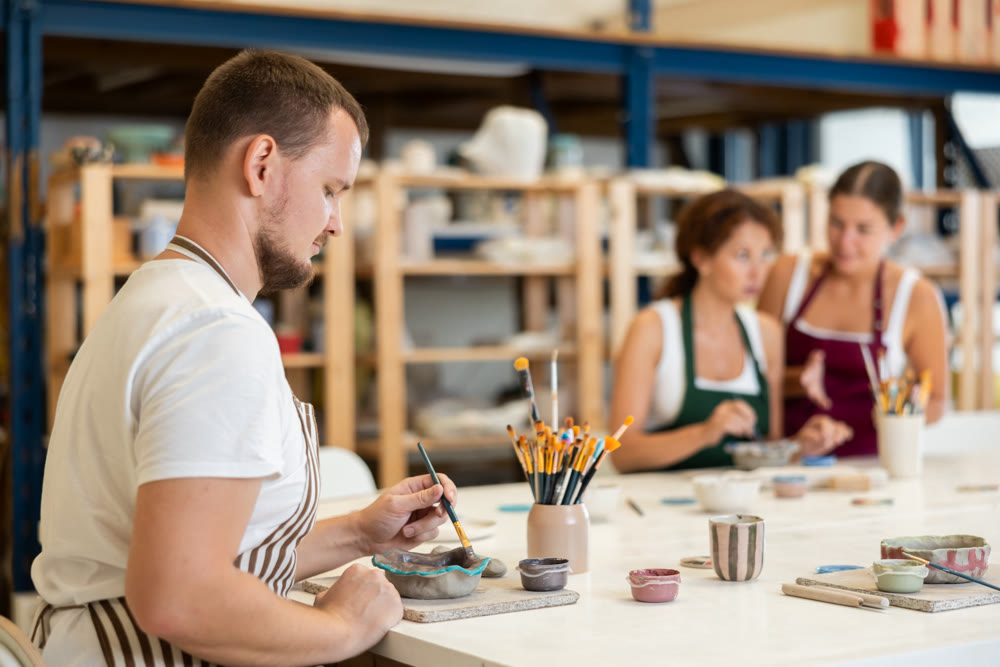 Male potter learns how to make designs on clay plate using brush and paint in pottery workshop