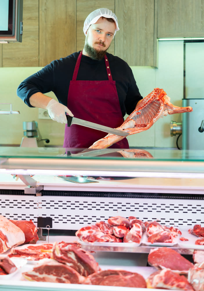 Skilled bearded butcher cutting fresh lamb carcass at counter