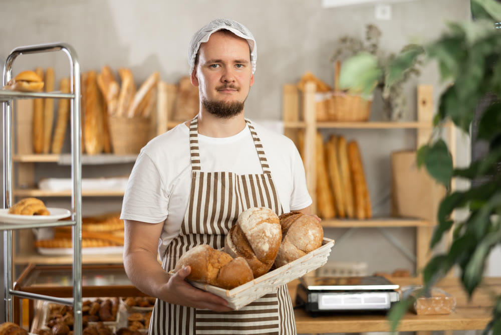 In sales area room of bakery, there is guy with loaf breads and baguetttes in wicker basket in hands