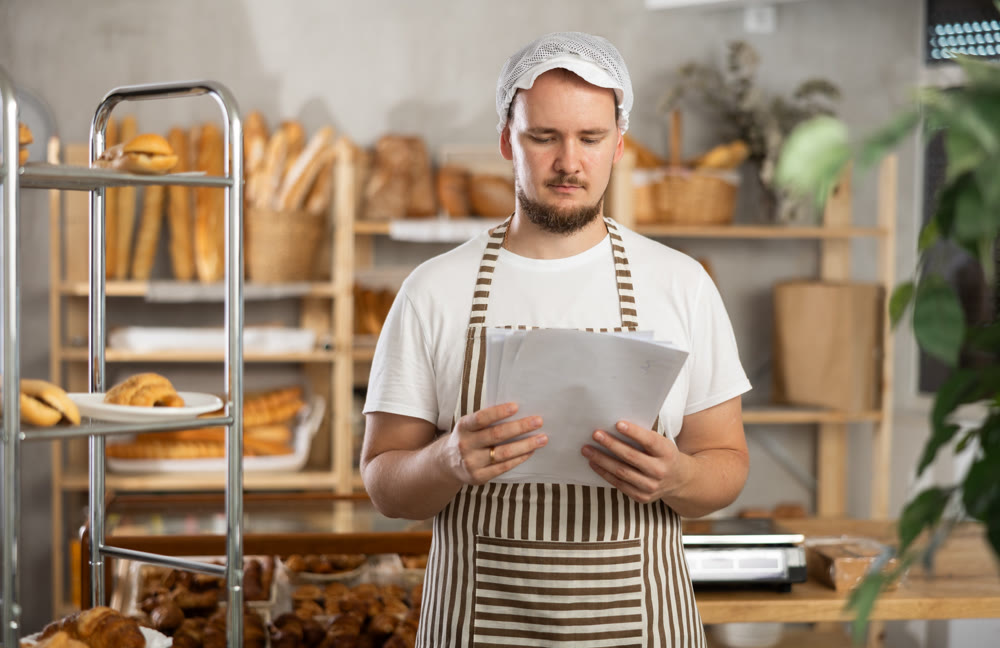 Guy running bakery is standing with papers in hands in middle of bakery