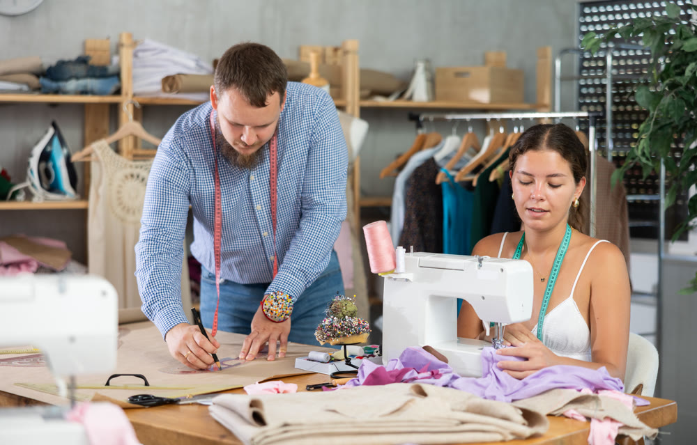 Female fashion designer sewing while colleague drafting pattern