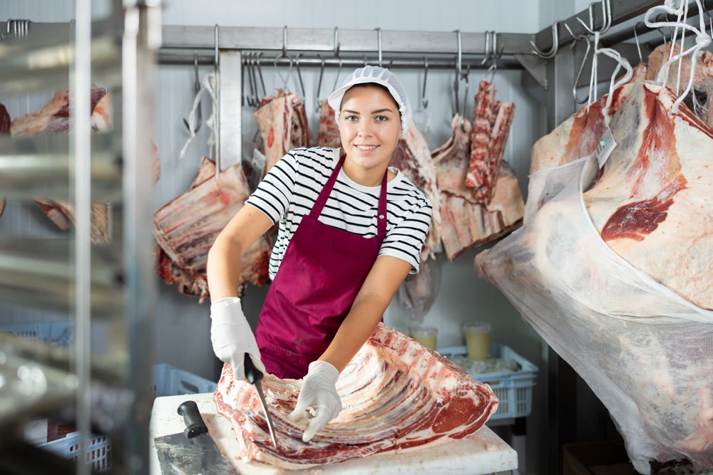 Positive female butcher cutting veal ribs in cold storage room of store