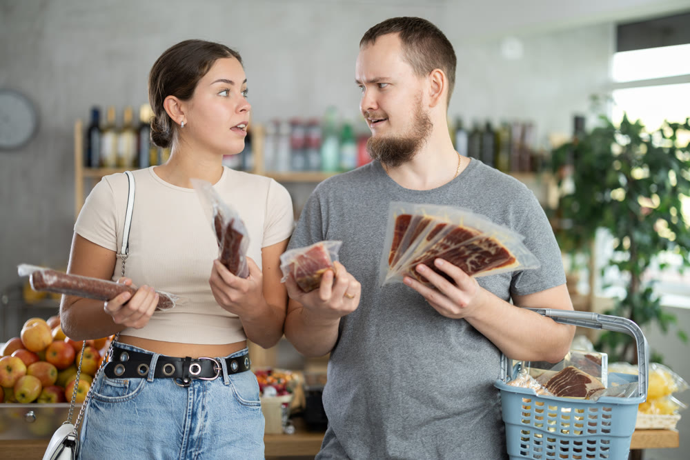 Satisfied male and female customers with dried sausage and jamon in hands shopping together in grocery store