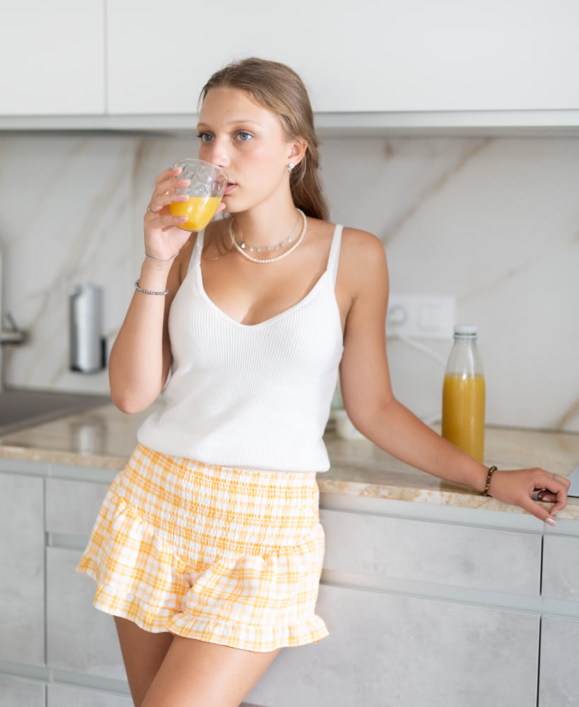 Young positive girl in casual clothes home T-shirt and shorts drinking juice or smoothie on kitchen