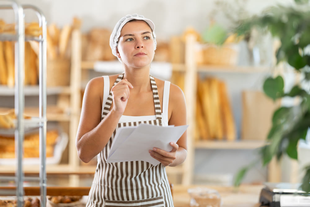 Thoughtful young woman holds papers standing in bakery