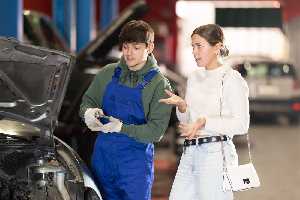 Mechanic advises young woman client on repairing under hood of car in car service station