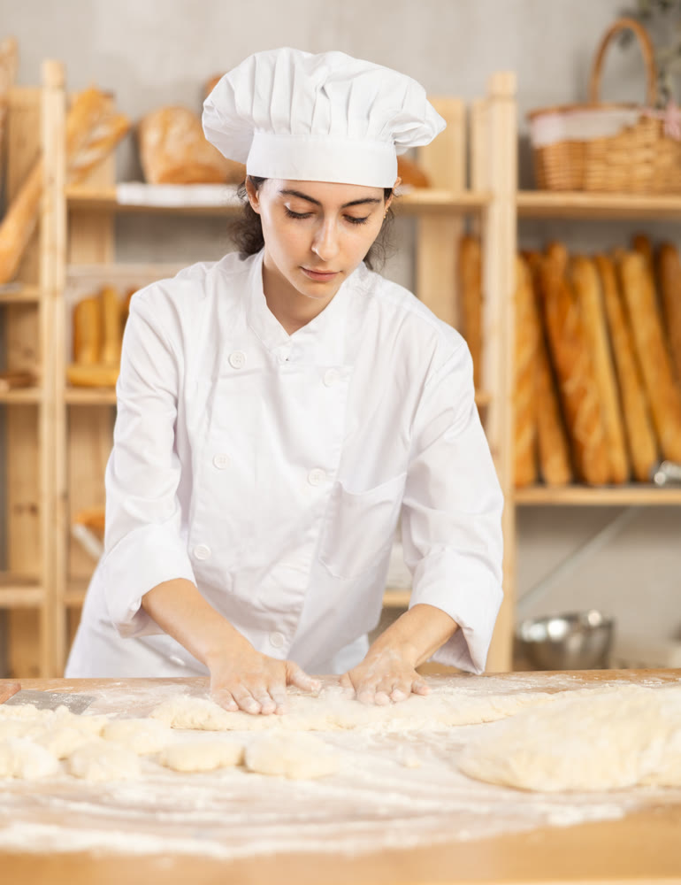 Young female baker kneading dough on table