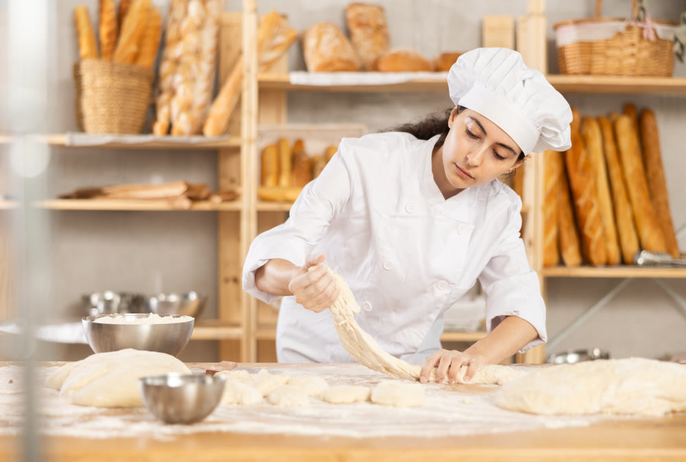 Young woman works as baker, mold baguette from dough, creates long loafs from dough.