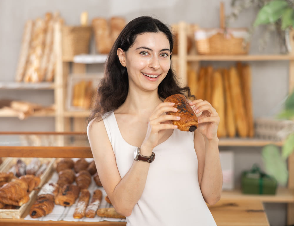 Young female customer stands with croissant in hands near window of bakery.