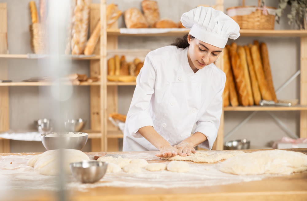 Young female baker kneading dough on table