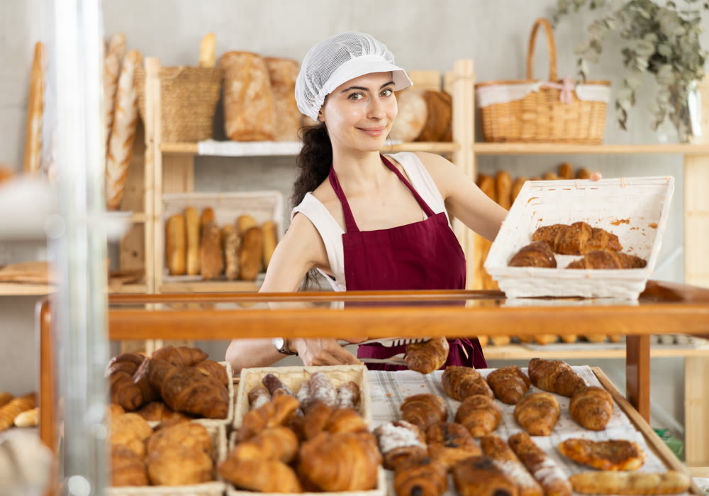 Young woman employee puts croissants in window, arranges display of goods at bakery.