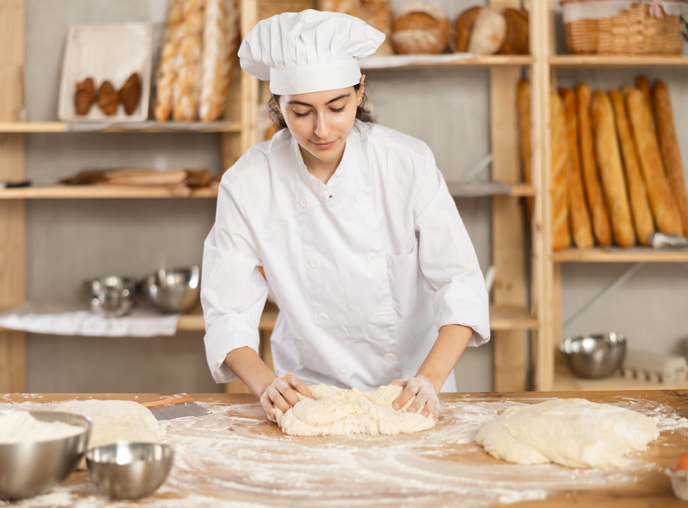 Young woman works in bakery as baker, kneads dough, works with flour.