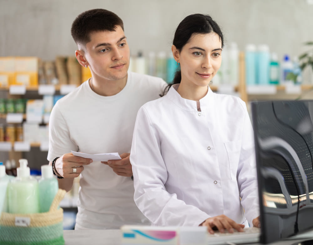 Male customer holding prescription searches for medicine in pharmacy. Female pharmacist offers to help find information on computer