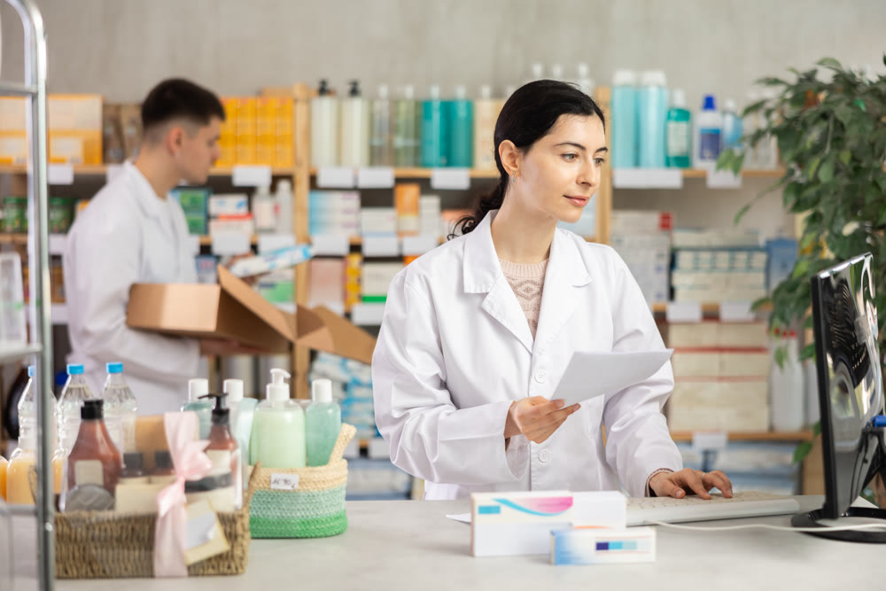 Female pharmacist in uniform looking for medicine in pharmacy using computer. In background, male pharmacist places