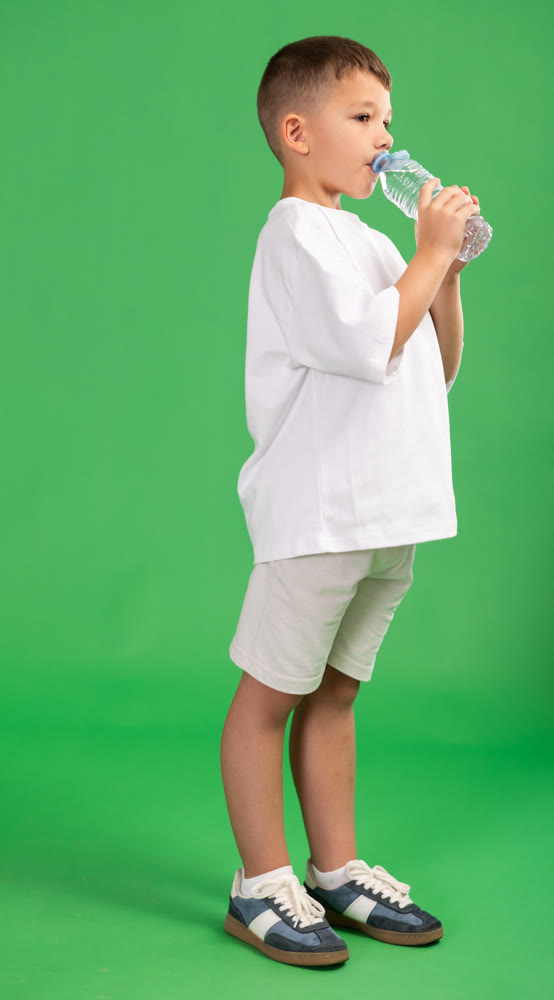 Boy in white clothes drinks water from a plastic bottle on green background