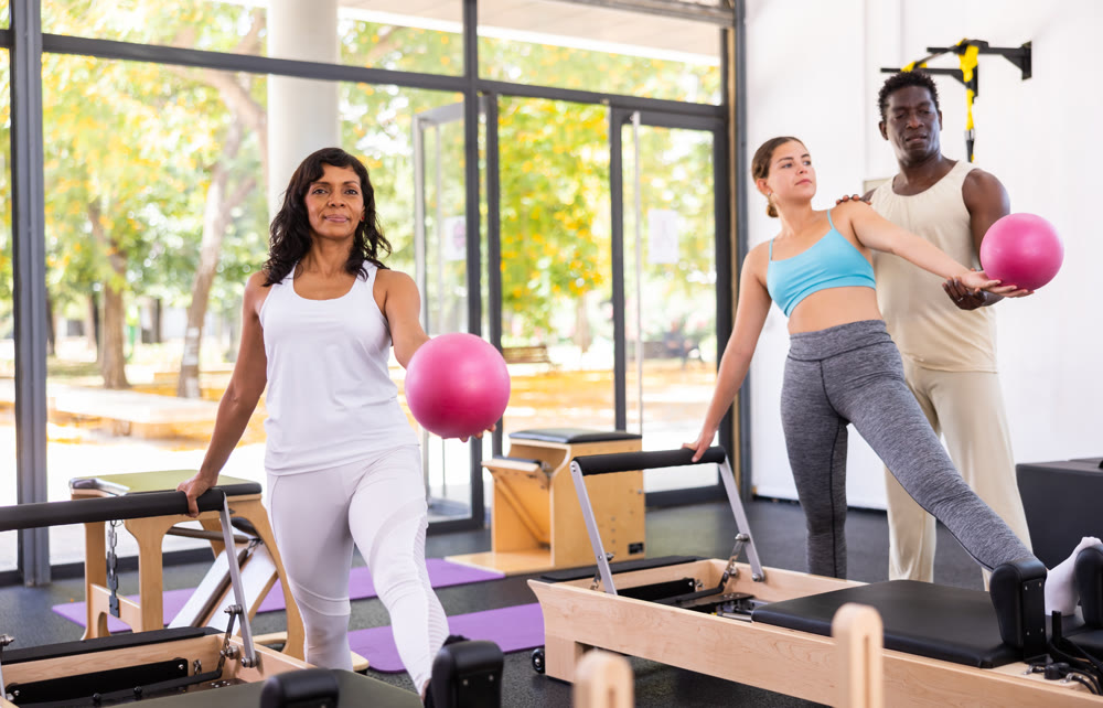 Attentive African American male trainer helping two positive women training with pilates bender ball