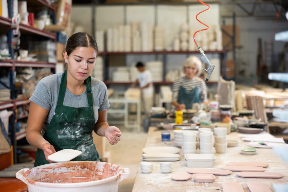 Young female worker in pottery factory lowers fired clay plates and cups into container with glaze
