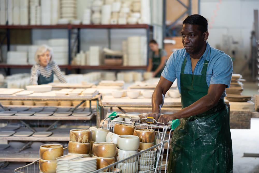 African american man in ceramics factory carries many different clay ceramic cups and plates in cart