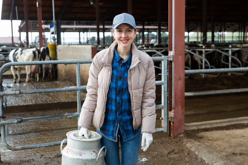 Female farmer standing near cowshed at dairy farm with milk churn