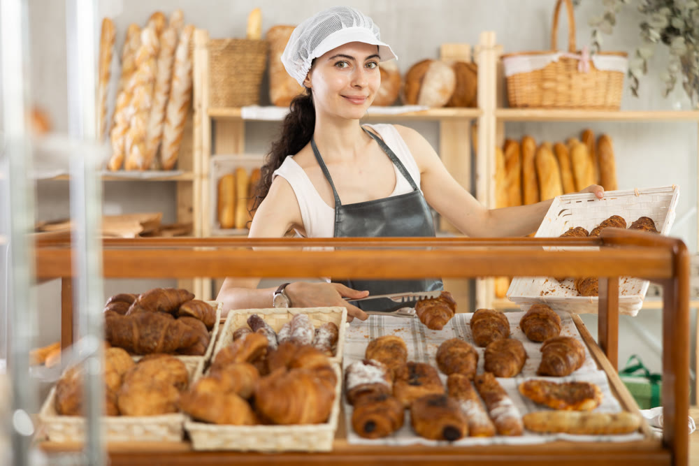 Young woman employee puts croissants in window, arranges display of goods at bakery.