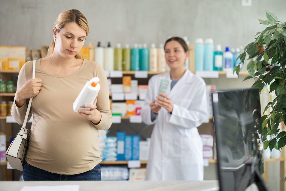 Pregnant woman choose diaper rash cream in pharmacy, pharmacist in background offer similar product