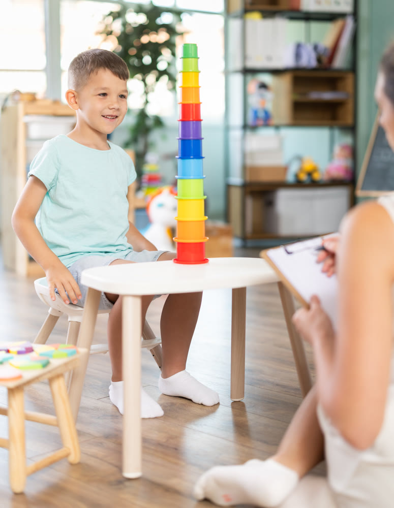 Boy plays with multi colored plastic cups, placing them inside each other. Psychologist helps and observes.