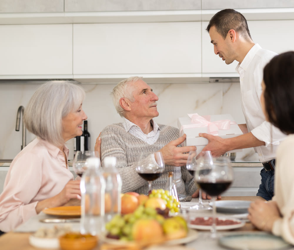Young guy giving present to old man while sitting around kitchen table