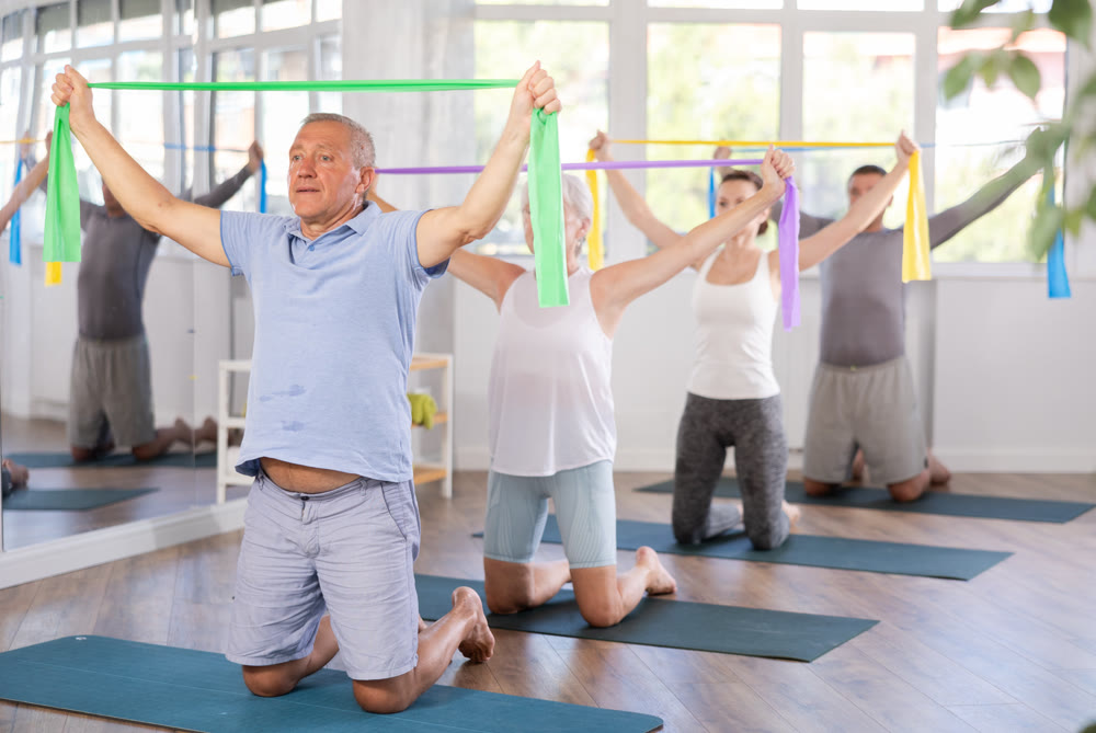 Group of mature people doing arm stretching exercises using resistance bands while kneeling on mats
