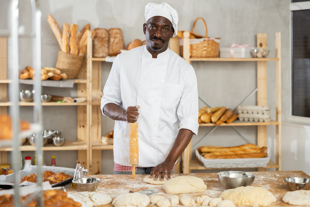 African baker rolling dough on floured table in bakery