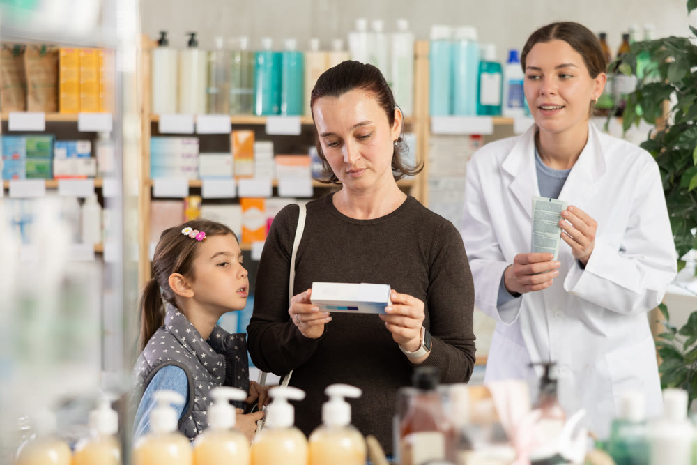Woman with schoolgirl daughter are busy choosing ointment at pharmacy.Pharmacist in background